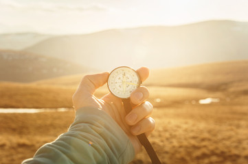a man's hand holds a hand-held compass against the backdrop of mountains and hills at sunset. The...