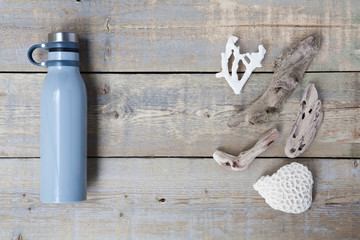 Blue stainless steel, reusable drink container flat lay on a light brown wooden table next to some driftwood, coral and a shell. With copy space. Horizontal.