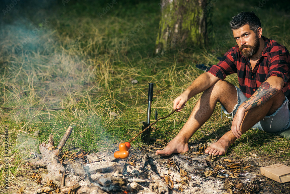 Wall mural Lone wanderer camping in woods. Guy frying sausages over fire. Barefoot man sitting on grass in forest