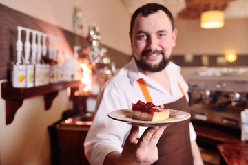 an attractive male confectioner in an apron holds a piece of mouth-watering cake with raspberries on the background of a cozy coffee house