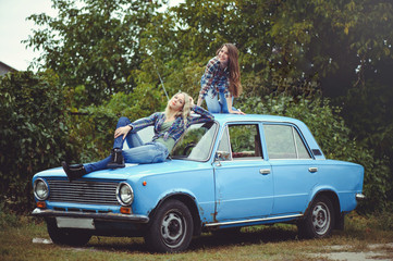 cheerful Attractive two young blonde girl and brunette posing on the hood of an old rusty car, dressed in jeans and shirts on a nature background