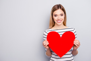 Portrait with copyspace, empty place for advertisement of attractive, charming, cheerful, cute, sweet girl with hairstyle having big red paper heart in arms isolated on grey background