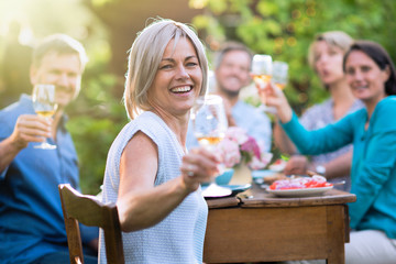 In summer. a group of friends in their forties gathered around a table in the garden to share a meal. They toast with their glasses of wine to the camera.