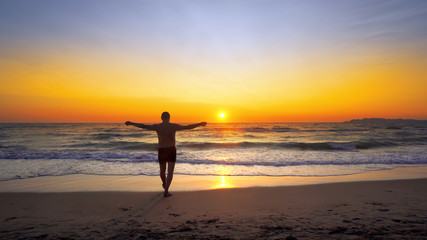 Fototapeta na wymiar Man with hands wide open in VICTORY pose walks on empty ocean beach into water with vibrant sunset sun at background, cinematic steadicam shot
