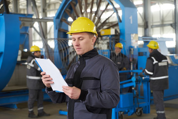 Portrait of factory worker holding paper clipboard. Young engineer in uniform and hardhat taking notes working at metal production factory, copy space