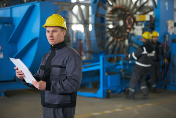 Young Worker reading paper documents while standing at factory with working machines on background. 