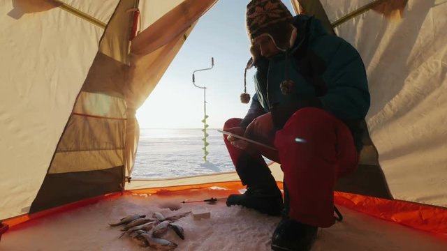 Man in fishing tent using tablet and waiting fish for bite on hole in winter