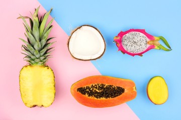 Tropical fruit flat lay with pineapple, dragon fruit, papaya, mango, and coconut on a pastel pink and blue background