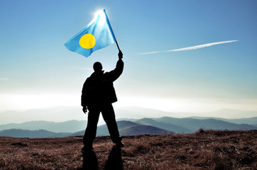 Successful silhouette man winner waving Palau flag on top of the mountain peak