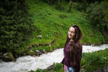 Young woman stands in national park on background of green hill with mountain river in summer day. Trendy girl looking away and smiling.