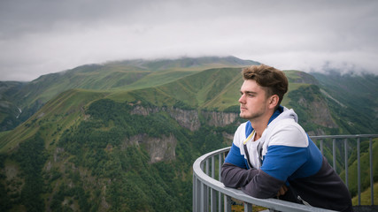 Young man stands at observation deck on background of beautiful green mountains and sky. Handsome guy have a rest before hiking.