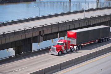 Classic red big rig semi truck with black semi trailer running on the multilevel overpass road over the river © vit