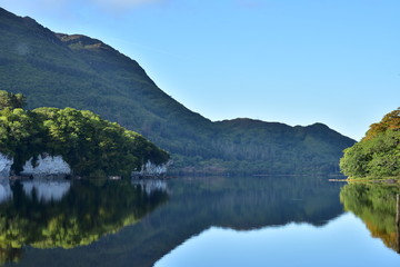 Fototapeta na wymiar Reflections of hills forests and rock formations on calm surface of Muckross Lake in Ireland.