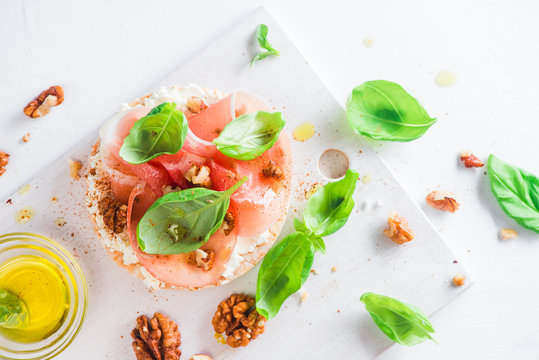 Crisp bread healthy snack with Parma ham, soft creme cheese, olive oil, fresh basil leaves, walnuts. Easy breakfast close-up on a white background with copy space.