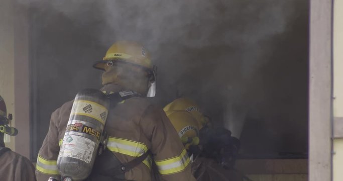 Slow-motion firefighters spraying water through a window of a burning house
