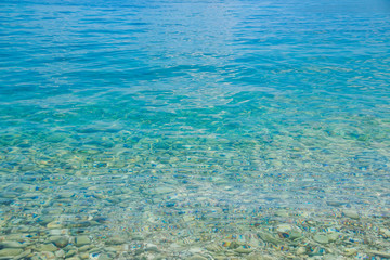 Fototapeta na wymiar Transparent shallow water with rocky bottom, fading away to deeper area at top.Crystal clear water with transparent surface shine under the bright summer sun. Colorful underwater stones.