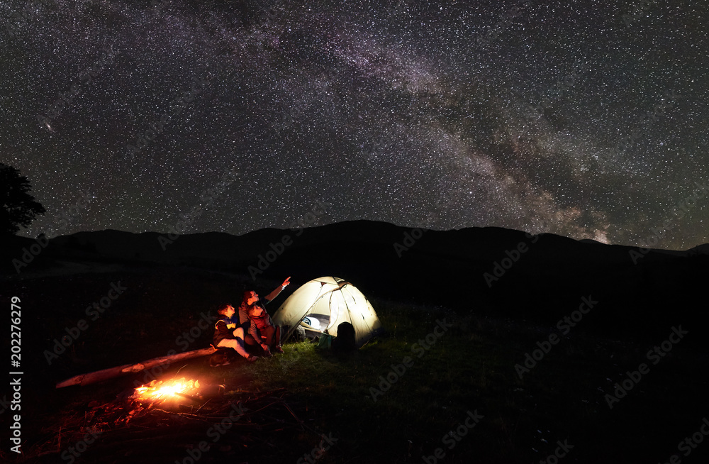 Wall mural Father and two children tourists resting at night camping in mountains, sitting on log beside campfire and glowing tent, looking at incredible beautiful starry sky and Milky way. Man pointing at sky