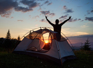 Silhouette of a tourist female standing with her hands raised to the sky on top of a mountain near campsite at sunrise. Through tent visible bright sun rises over the mountains. Rear view