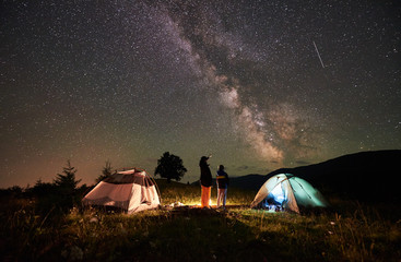 Back view mother and son tourists resting at camping in mountains, standing beside campfire and two tents, looking at night sky full of stars and Milky way, enjoying night scene. Woman pointing at sky