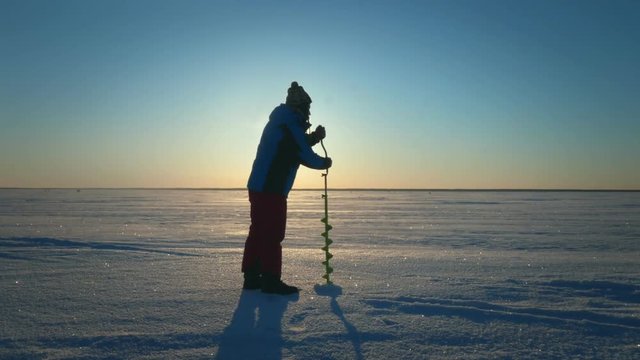 Fisherman drilling hole in ice lake for winter fishing on skyline background
