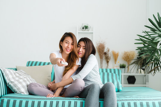 LGBT lesbian women couple moments happiness. Lesbian women couple together indoors concept. Friends young smiling women at home sitting on the couch and watching tv, She is holding a remote control.