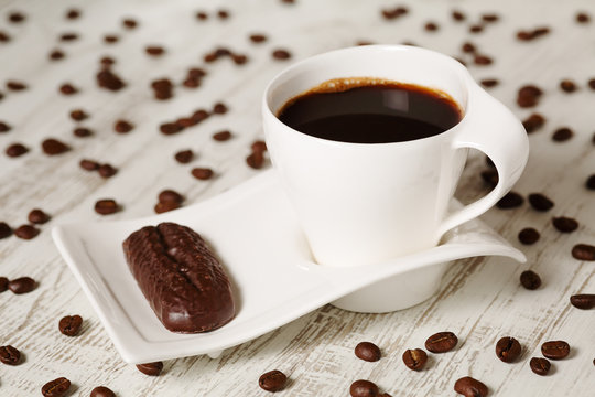 Morning coffee in white ceramic cup with wave shaped saucer and chocolate biscuit on white wooden rustic table with scattered coffee beans