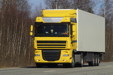 Fototapeta na wymiar Transportation logistics - yellow truck with white semi-trailer on asphalt road in spring against blue sky forest, front view