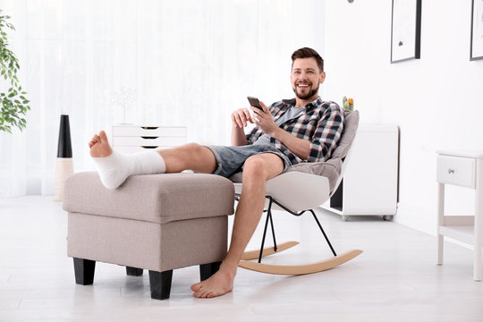 Man with broken leg in cast using mobile phone while sitting in armchair at home