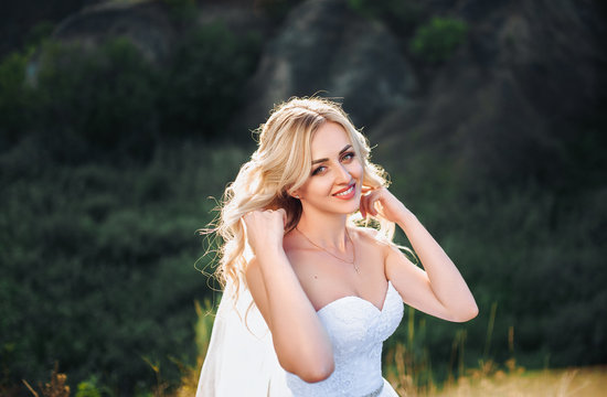 A beautiful bride plays with her curly hair against the backdrop of hills and sunny nature. Portrait of a bride with sunlight in the evening. Smiling Bride.