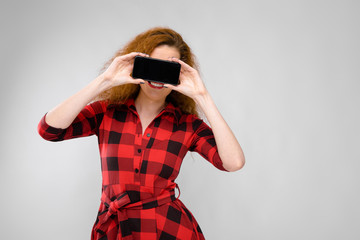 Portrait of beautiful redhead happy young woman in checkered dress smiling holding mobile phone in front of her face on gray background