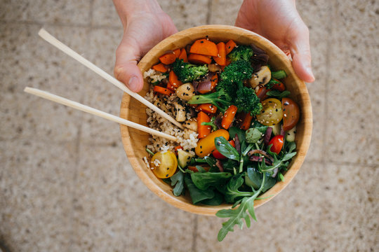 vegan or vegeterian man or woman holds up wooden coconut buddha bowl full of healthy greens, vegetables, grains and chopsticks. Concept lifestyle choice and mindset food blog