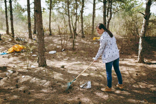Young woman in casual clothes cleaning rubbish using rake for garbage collection in littered park or forest. Problem of environmental pollution. Stop nature garbage, environment protection concept.