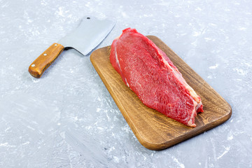 fresh large piece of beef on a wooden cutting board with kitchen ax. Top view on gray concrete...