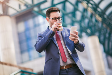 Businessman using mobile phone outside of office buildings in the background. Young caucasian man holding smartphone for business work and drink coffee to go.