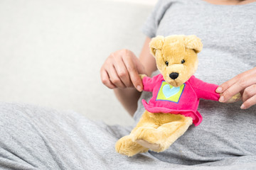 Happy pregnant woman hand holding teddy bear doll on her belly while sitting on sofa in living room at home.