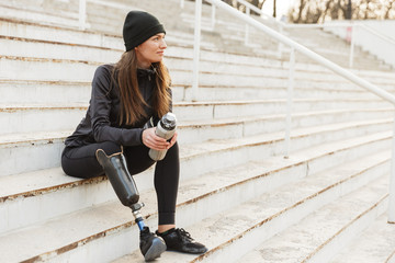 Portrait of attractive handicapped woman in black tracksuit with prosthetic leg sitting at the street stairs, and holding thermos cup