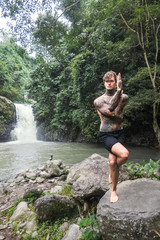 tattooed man practicing yoga on rock with aling-aling waterfall and green plants on background, Bali, Indonesia