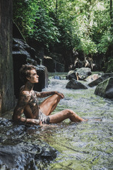 side view of tattooed man resting on rocks with water and green plants around, bali, indonesia