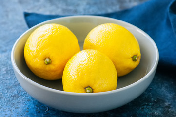 Close up of three ripe yellow lemons in a grey bowl on a blue stone background.