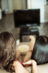 Fototapeta premium Back view photo of two girlfriends who are sitting on the couch watching TV.