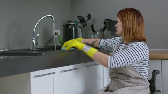 Medium shot of independent woman in wheelchair turning on water faucet in kitchen and washing dishes