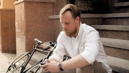 Fototapeta na wymiar Portrait of stylish hipster man with beard using smartphone while sitting on street