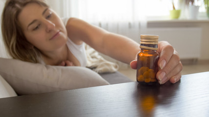 Closeup photo of young ill woman lying on sofa and taking bottle with pills from bedside table