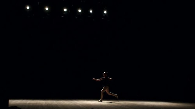 Girl ballerina doing horizontal twine in the air on stage on black background, slow motion. Girl doing gymnastic flip. Acrobatic flip. Ballet. Dancer.