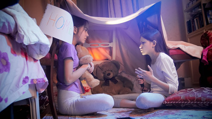 Obraz na płótnie Canvas Two teenage girls in pajamas sitting with flashlight in tepee tent in bedroom at night