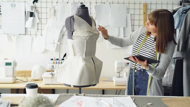 Young fashion designing entrepreneur is working with tablet while checking clothing pinned to tailoring dummy. Modern technology in clothes manufacturing concept.