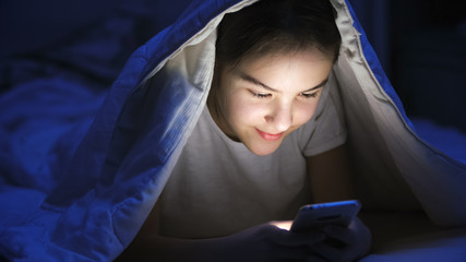Portrait of teenage girl typing message on smartphone under blanket at night