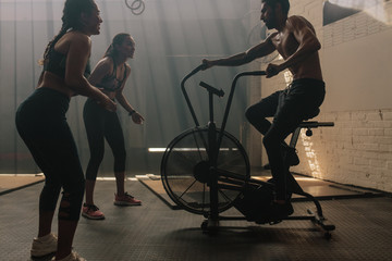 Fototapeta na wymiar Two women motivating man exercising on air bike in gym