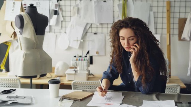 Professional fashion designer is talking on mobile phone and drawing clothing sketch with pencil at table in tailoring shop. Everyday work and communication in workplace concept.