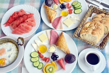 Turkish breakfast, top view, sliced bread in basket, various cheese, ham, puff, butter, jam, olive, tomatoes, kiwi, orange  and cucumber in white plate, watermelon and fried eggs and black coffee 